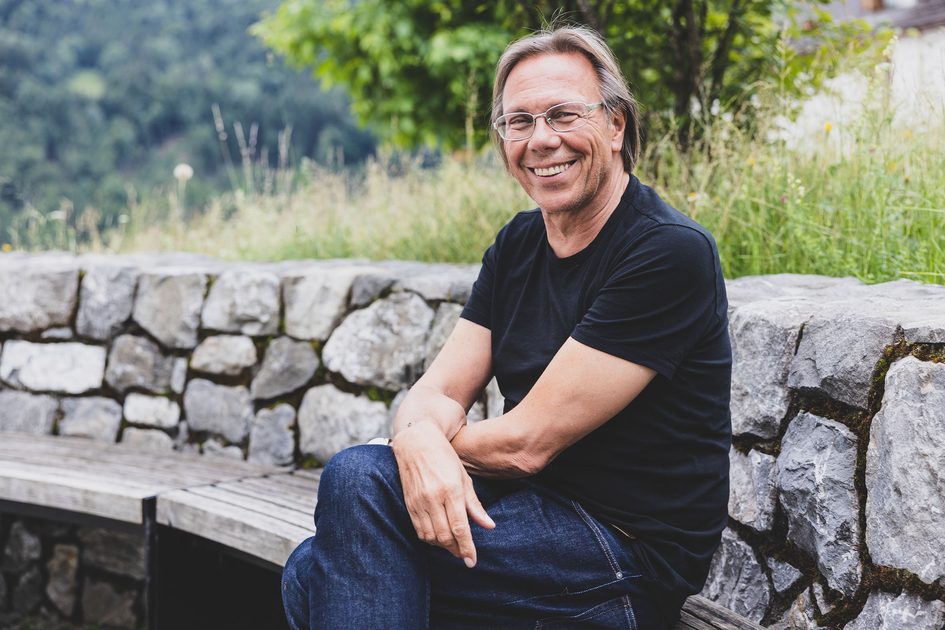 Ein lächelnder Mann mit Brille, schwarzem T-Shirt und Jeans sitzt auf einer gebogenen Holzbank vor einer niedrigen Steinmauer in einer grünen Parklandschaft