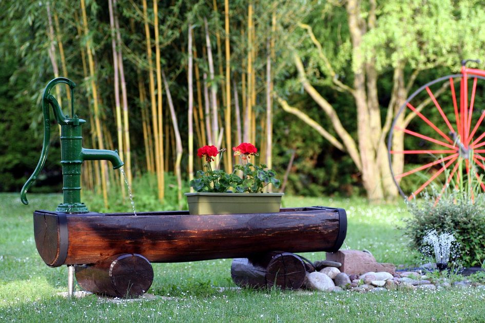 Gartenbrunnen aus einem Baumstamm mit Handschwengelpumpe und Geranien im Blumentopf