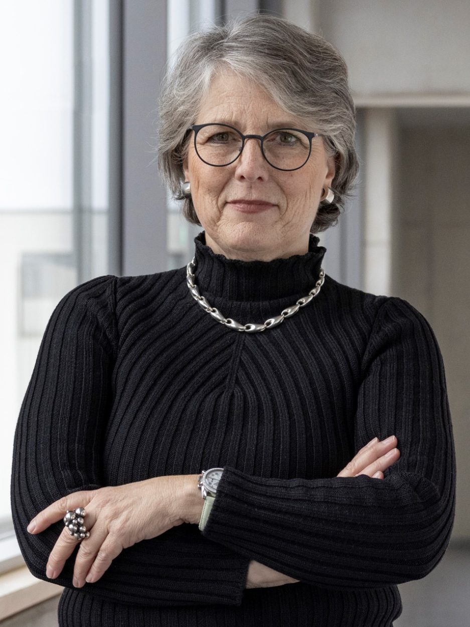 Eine Frau steht neben einem großen Fenster. Sie trägt eine Brille und lächelt.