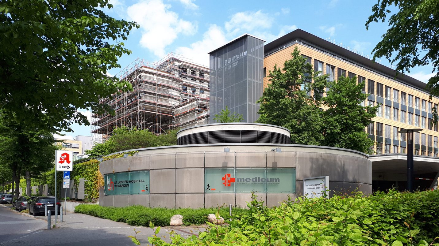 Das Bild zeigt eine Außenansicht des St. Josefs-Hospitals Wiesbaden. Im Vordergrund befindet sich ein rundes, modernes Gebäude mit der Beschriftung "medicum – Facharztzentrum Wiesbaden". Das Gebäude hat eine metallisch-silberne Fassade mit begrünten Wänden. Im Hintergrund sieht man das Hauptgebäude des Krankenhauses, ein mehrstöckiges Gebäude mit einer Holz-Glas-Fassade. Daneben befindet sich ein weiteres Gebäude, das von einem Gerüst umgeben ist, was auf Bau- oder Renovierungsarbeiten hindeutet. Auf der linken Seite des Bildes steht ein Verkehrs- bzw. Hinweisschild mit einem roten Krankenhaus-Symbol, das den Weg zu einer Fußgängerüberquerung und einem Parkplatz weist. Die Umgebung ist von Bäumen und grünen Sträuchern gesäumt, und der Himmel ist blau mit wenigen Wolken.