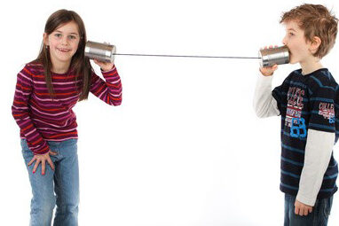 Zwei Kinder mit Dosentelefon, auf der linken Seite hält ein Mädchen mit langen, braunen Haaren die Metalldose ans Ohr und schaut frontal in die Kamera. Sie trägt eine Jeanshose und einen gestreiften Pullover in lila, rot und rosa. Der Junge auf der rechten Seite des Bildes ist von der Seite zu sehen, er hält sich die Dose an den Mund. Er hat blonde Haare, trägt eine dunklere Jeans und ein schwarzes T-Shirt mit blauen Streifen und Aufdruck. Darunter trägt er ein weißes Oberteil mit langen Armen. Die Dosen sind durch eine Schnur miteinander verbunden.