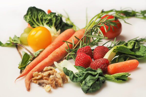 Nahrungsmittel wie Karotten, gelbe und rote Tomate, Himbeeren, Mangoldblätter, Walnüsse und ein Zweig Rosmarin liegen hübsch drapiert auf einer weißen Oberfläche.