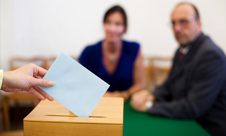 Eine Hand wirft einen Umschlag uin eine Wahlurne. Im Hintergruind zwei Men
