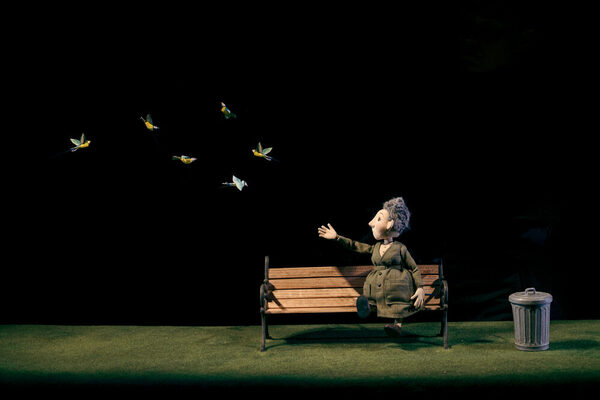 Una bambola a forma di donna anziana con un cappotto di lana verde siede su una panchina del parco e allunga il braccio destro. Nel punto in cui punta il braccio, sei uccelli giallo-blu volano verso di lei. Lo sfondo è nero. La donna sembra voler salutare gli uccelli.