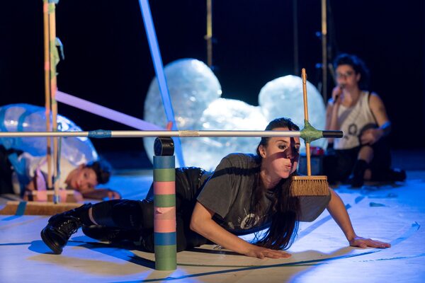 Una giovane donna è sdraiata su un palcoscenico in primo piano, di fronte a lei una torre fatta di rotoli di nastro adesivo, su cui giace trasversalmente un manico di scopa. Una piccola scopa è attaccata al lato della scopa. La donna vi soffia contro.