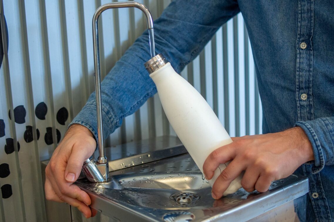 Eine Person im blauen Jeanshemd füllt eine weiße Wasserflasche am Wasserhahn auf.