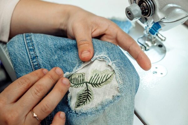 Foto von oben von einer Jeans mit einem Loch, zu sehen sind Hände die einen Flicken einbringen und Teile einer Nähmaschine. Der Flicken ist weiß und ist mit grünen Blättern bestickt.
