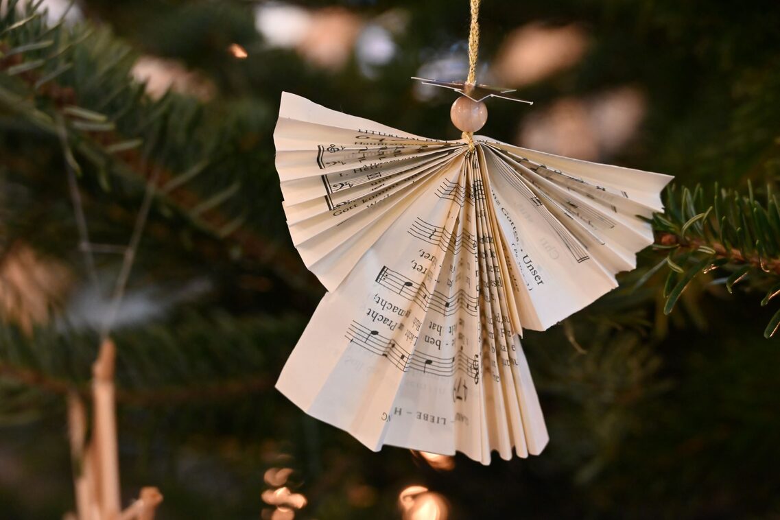 Weihnachtlicher Engel aus gefaltetem Notenpapier mit Holzperle als Kopf, hängend an einem Tannenzweig.