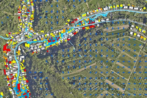 Kartenansicht eines Gebiets mit bunten Symbolen, die verschiedene Gebäudetypen (rot, gelb, blau) und Bäume (blaue Symbole) darstellen, flankiert von einem Fluss und umgeben von landwirtschaftlichen Flächen.