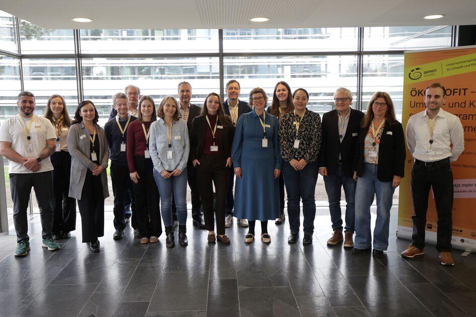 Gruppe von Menschen mit Namensschildern steht in einem hellen Raum vor großen Fenstern und einem gelben Banner.