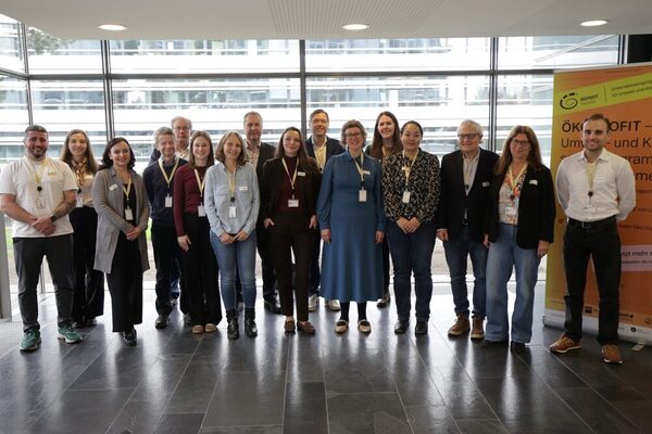 Gruppe von Menschen mit Namensschildern steht in einem hellen Raum vor großen Fenstern und einem gelben Banner.