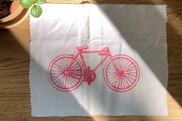 Ein weißes Stoffrechteck liegt auf einem Holztisch, es ist in roter Farbe mit einem Fahrrad bedruckt.