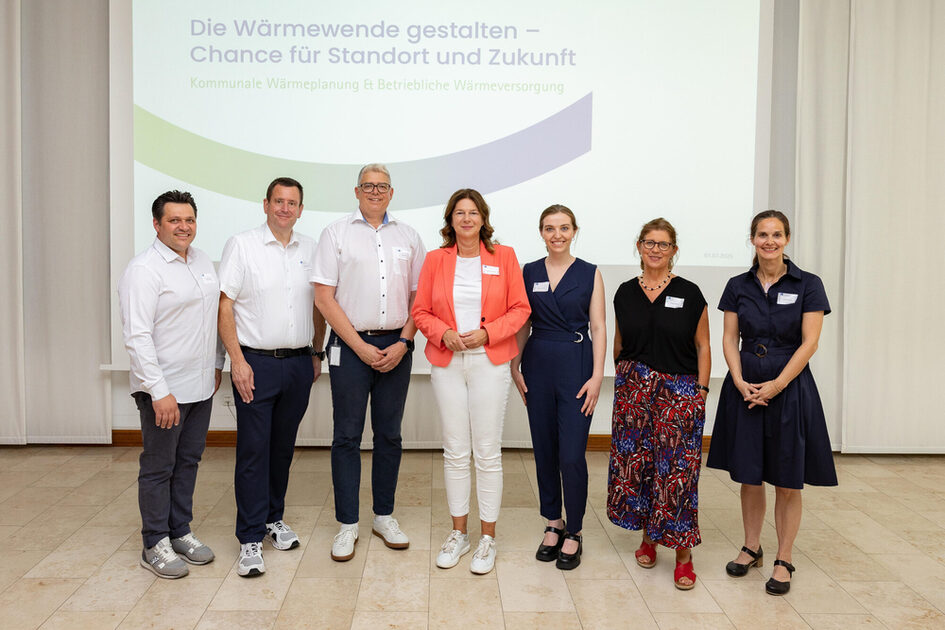 Sieben Personen stehen vor einer Leinwand mit dem Titel 'Die Wärmewende gestalten – Chance für Standort und Zukunft' bei einer Veranstaltung zur kommunalen Wärmeplanung und betrieblichen Wärmeversorgung.