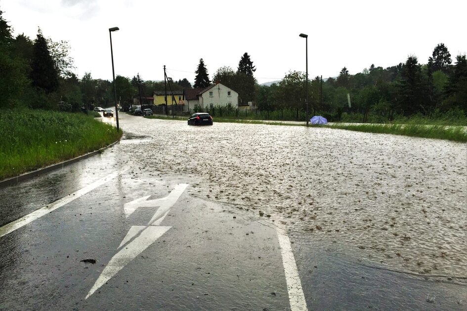 O stradă inundată în ploaie, două mașini pot fi văzute în apă pe drum, o umbrelă purpurie se află pe marginea drumului în fundal.