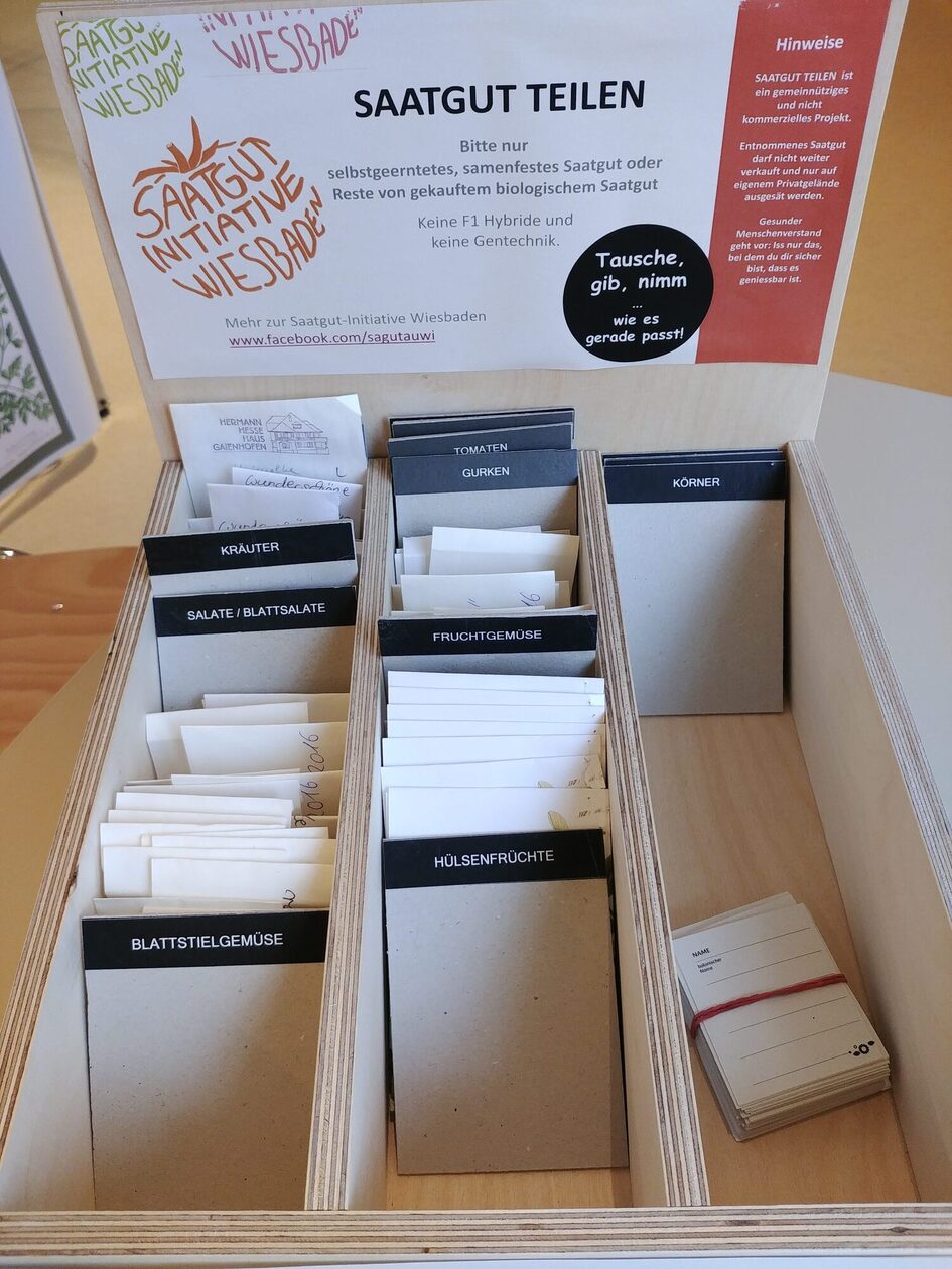 Holzkasten mit Karten zum Teilen von Saatgut, kategorisiert in verschiedene Gruppen wie Gemüse, Kräuter und Hölzer. Hinweis zur Saatgut-Initiative für nachhaltigen Anbau.