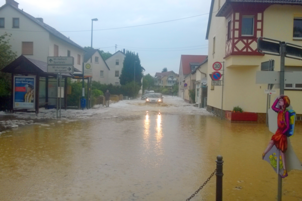Überflutete Straße mit stehendem Wasser, Häuser, Bushaltestelle und ein Auto im Hintergrund. Der Himmel ist bewölkt.