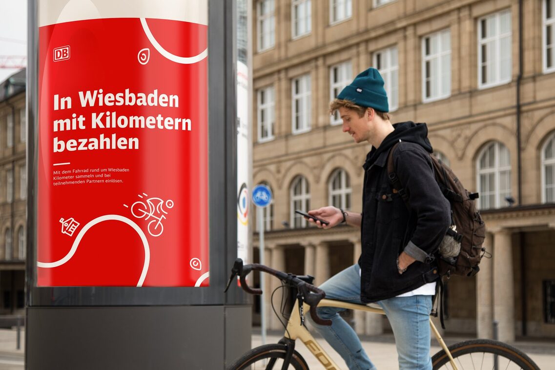 Ein junger Mann in lässiger Kleidung mit einem Rucksack steht an einem Fahrrad und schaut auf sein Smartphone. Im Hintergrund ist ein Werbeposter zu sehen, das für das Rad Programm in Wiesbaden wirbt.