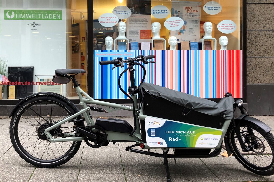 Lastenrad in grauer Farbe mit einem Werbeaufkleber für Verleihservice, vor einem Geschäft mit bunten Streifen im Hintergrund.