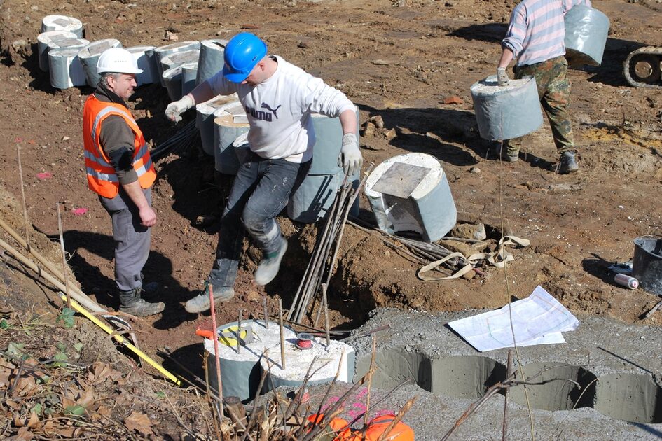 Bauarbeiter in Warnwesten und Helmen arbeiten auf einer Baustelle mit Betonfundamenten. Einer der Arbeiter hebt Eisenstangen, während ein anderer ihn beobachtet. Auf dem Boden liegen Betonformen und Bauunterlagen.