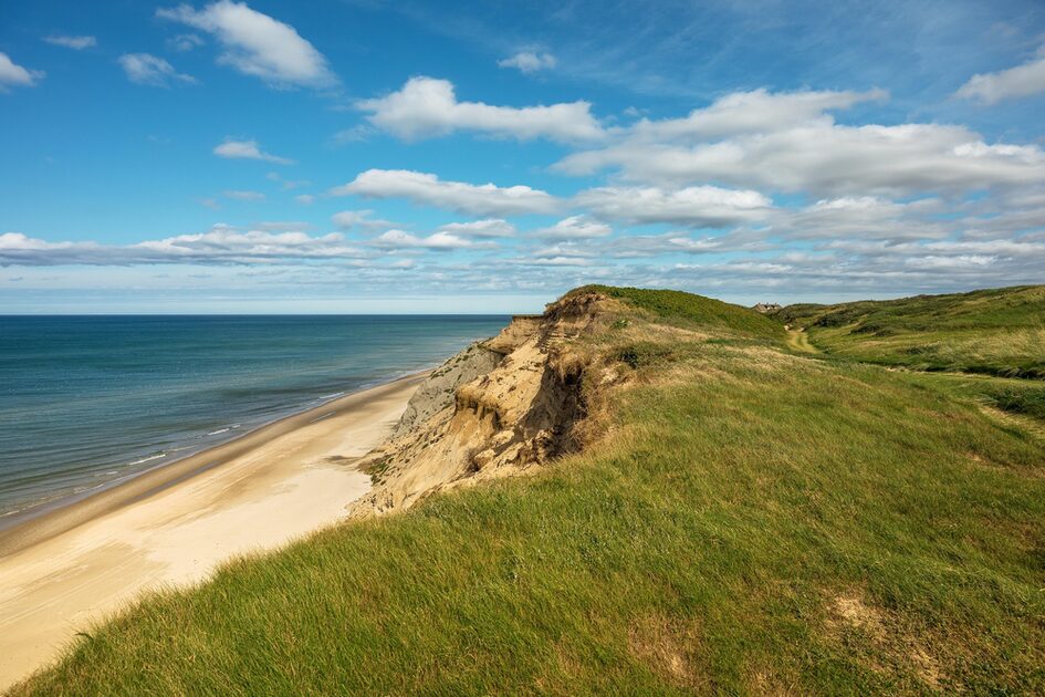 Grüne Dünen mit Blick auf einen sandigen Strand und ruhiges Meer unter blauem Himmel mit Wolken
