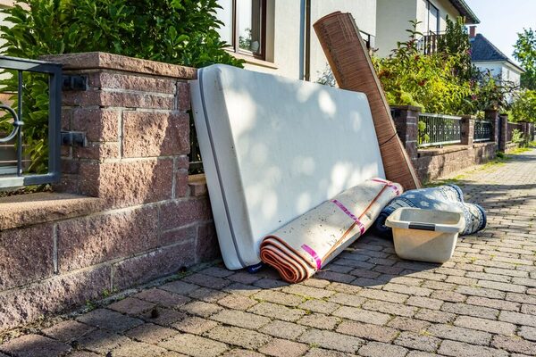 Bulky waste lies on a paved sidewalk. A mattress is leaning against the fence, rolled carpets and a white laundry basket lie in front of it.