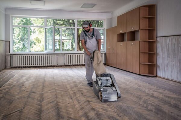 Mann schleift mit Schutzbrille und Gehörschutz den Holzboden in einem leeren Raum mit großen Fenstern