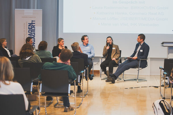 eine Gesprächsrunde sitzt vor einer Leinwand, eine Frau spricht in eine mikrofon, die anderen hören zu, vor einem gefüllten Saal.