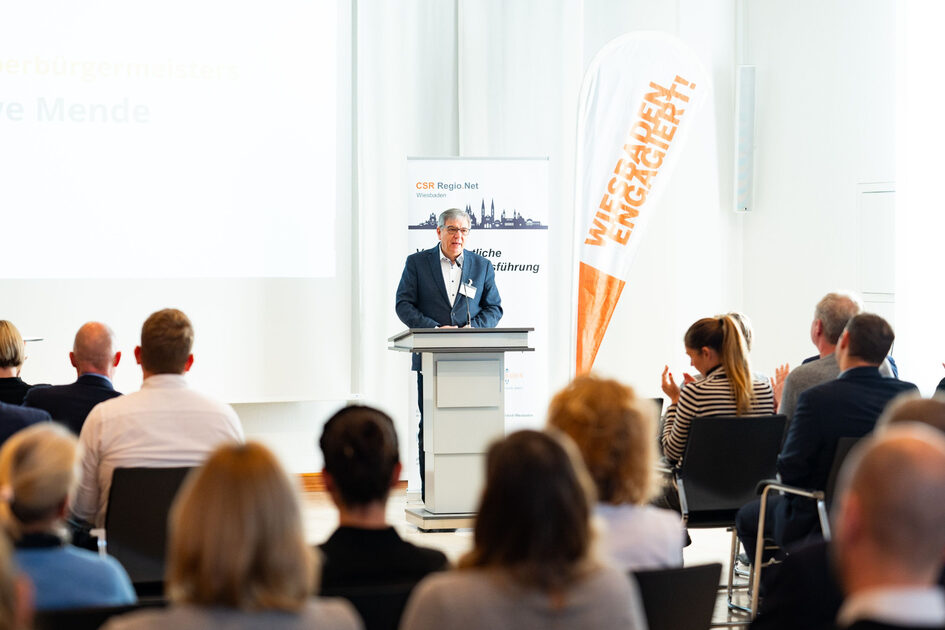Vor einem sitzenden Plenum spricht an einem Stehpult ein Mann. Es ist Oberbürgermeister Gert-UWe Mende. Im Hintergrund ist eine Beachflag. Auf dieser steht "Wiesbaden Engagiert!".