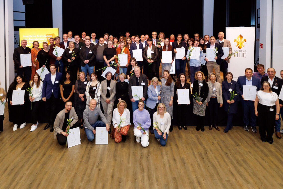 Viele Menschen stehen vor und auf einer Bühne, einige knien. Viele haben weiße Urkunden in der Hand, oder einzelne hohe Blumen.