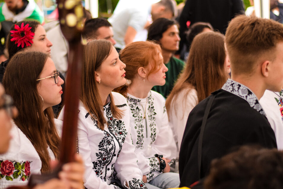 Viele junge Menschen sitzen und hören etwas zu. Die Mädchen tragen weiße, bestickte Blusen.
