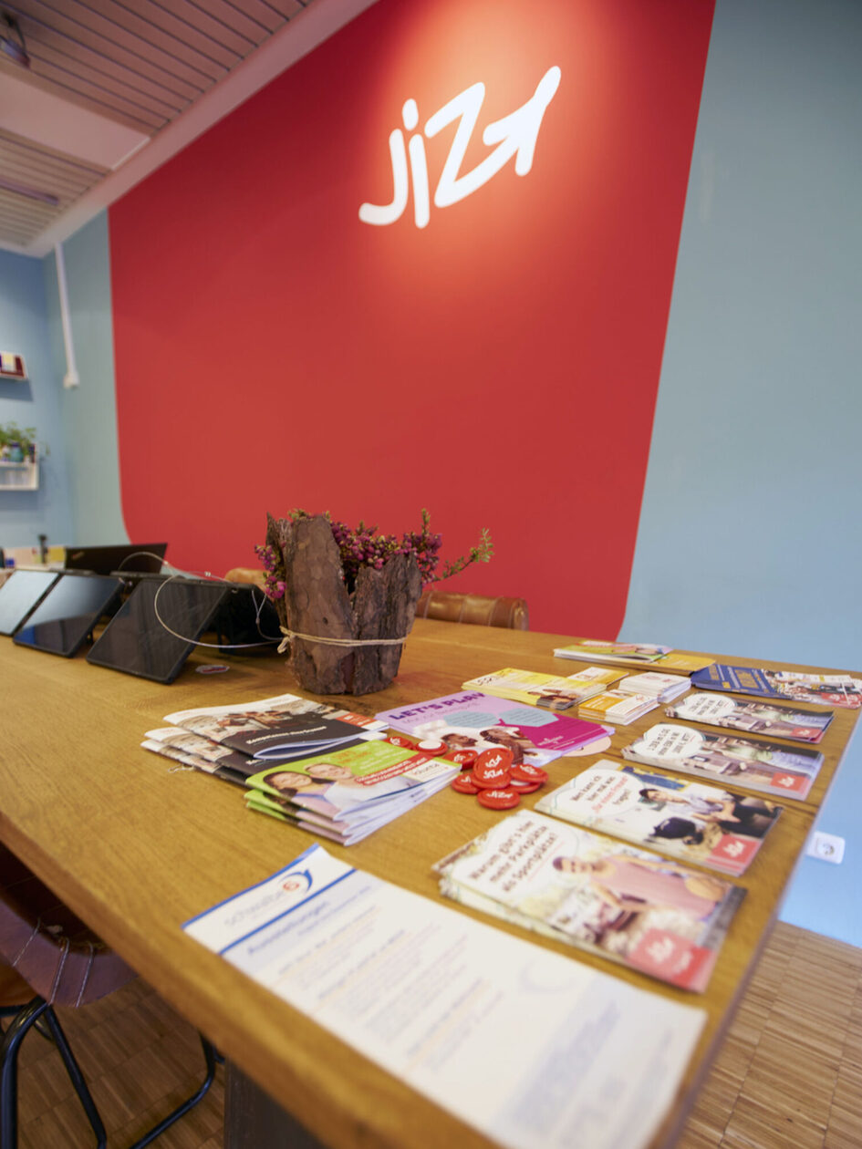 Auf einem Holztisch liegen verschiedene Flyer, im Hintergrund steht auf einer roten Wand "JIZ".