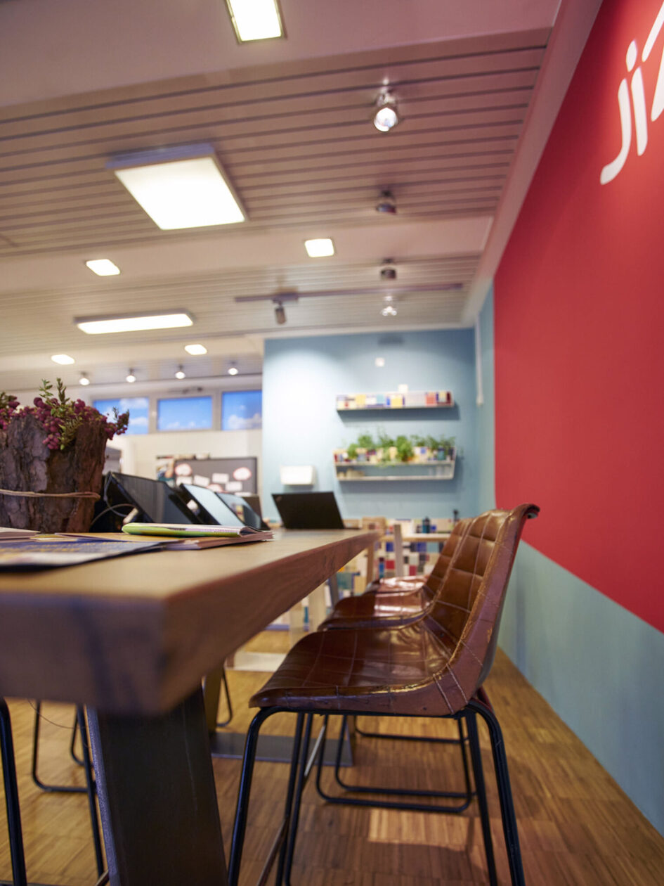 Im Vordergrund ist die Ecke einer Zischplatte zu sehen und eine Reihe Stühle. Auf einer roten Wand steht "JIZ".