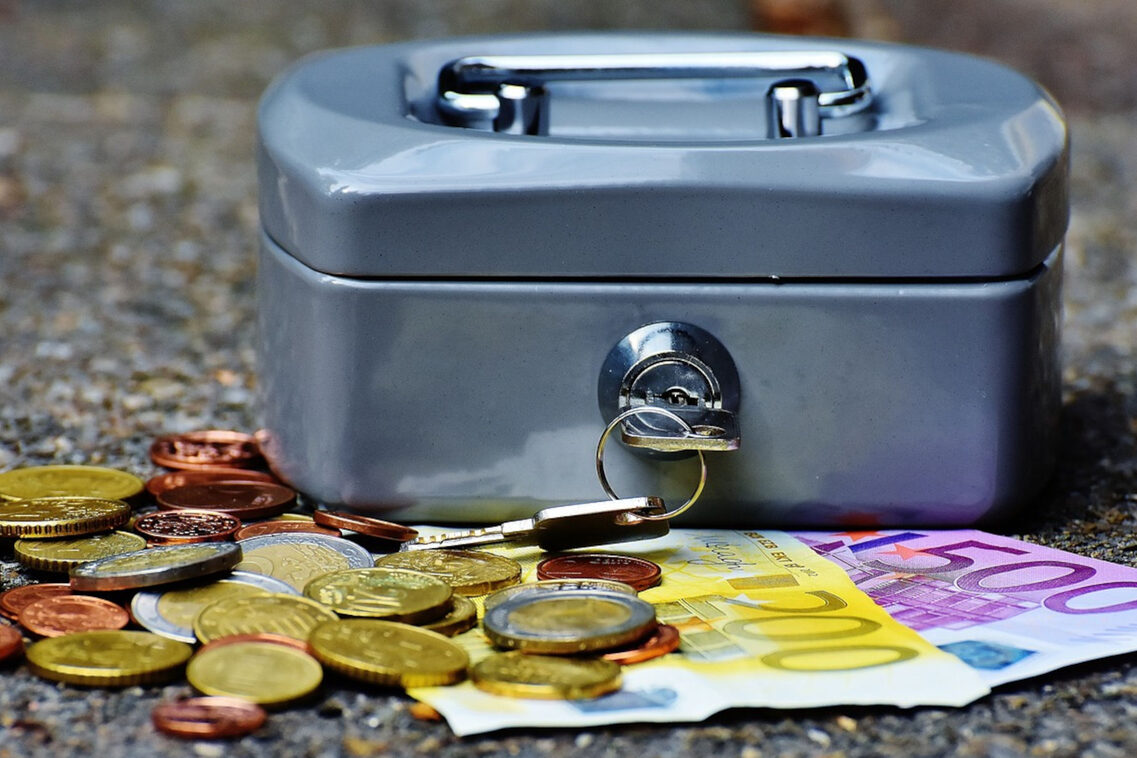 Eine graue Geldkassette steht auf dem Boden, daneben liegen Euromünzen und Euroscheine.