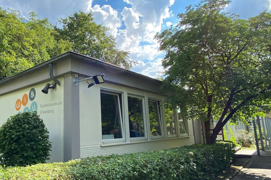 Ein weißes, einstöckiges Gebäude mit flachem Dach, inmitten von grünen Bäumen und Hecken. Auf der Seite steht "Stadtteilzentrum Gräselberg".