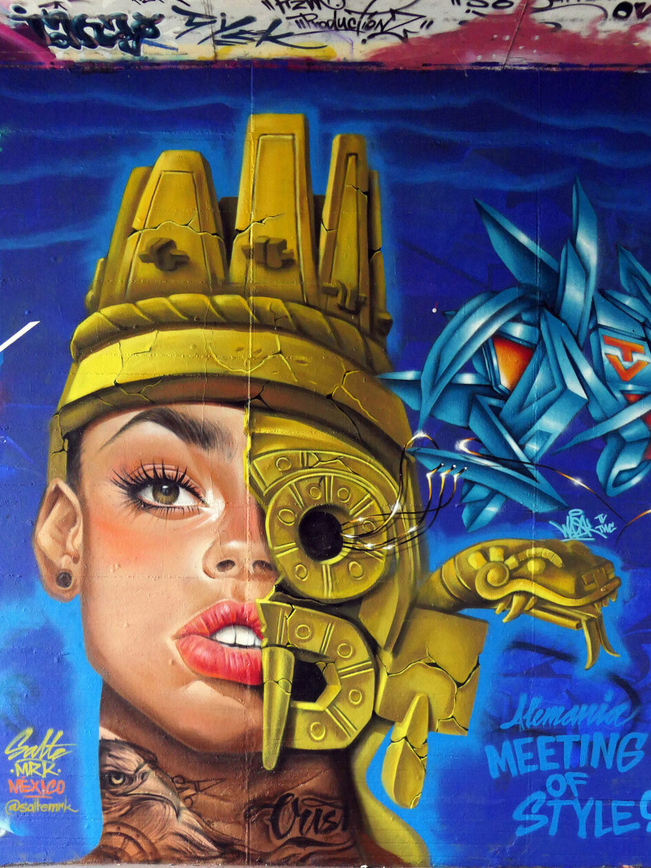 Das Graffiti zeigt einen Frauenkopf, der zur Hälfte mit Metallelementen bedeckt ist, auf dem Kopf sitzt eine Krone, vor blauem Grund.