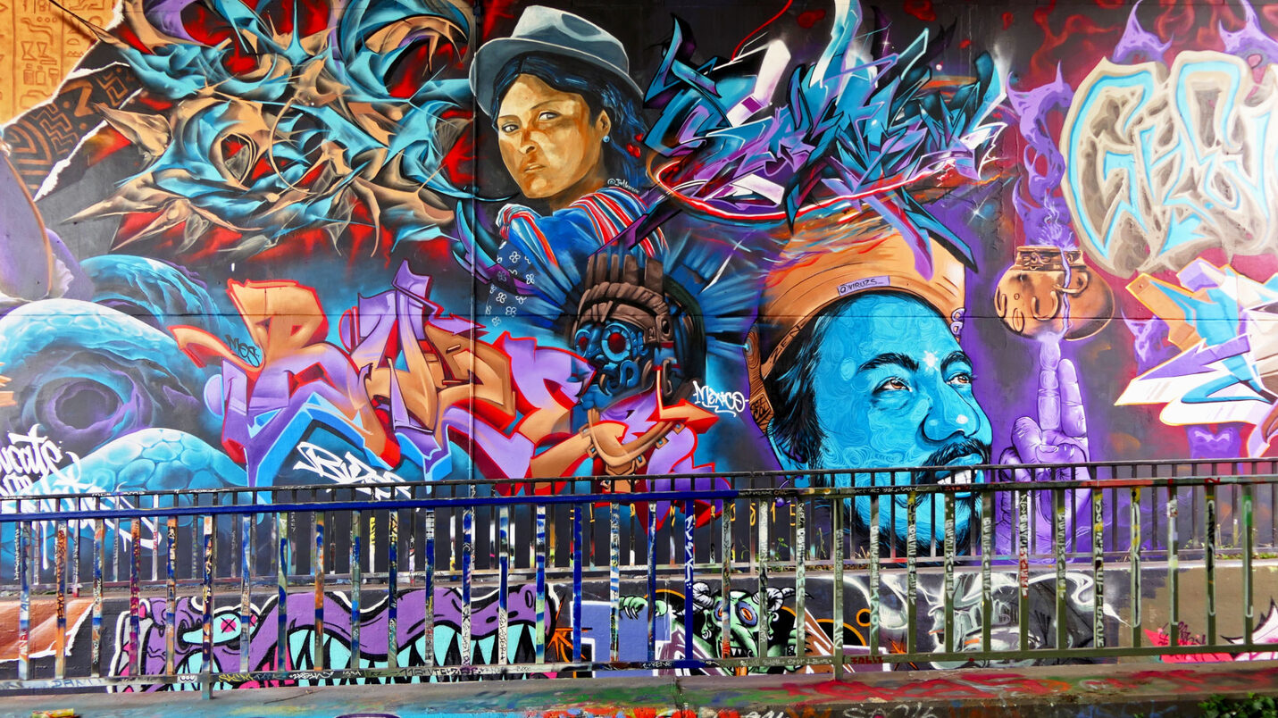 Buntes Graffiti, es sind verschiedene Köpfe zu sehen, Schriftzüge in violett, orange und blau.