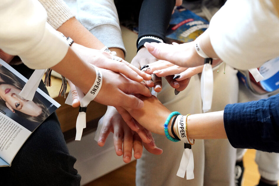 Mehrere Hände von jungen Menschen greifen zusammen in der Mitte. Sie tragen Armbänder, auf ihnen steht "Youth happens".