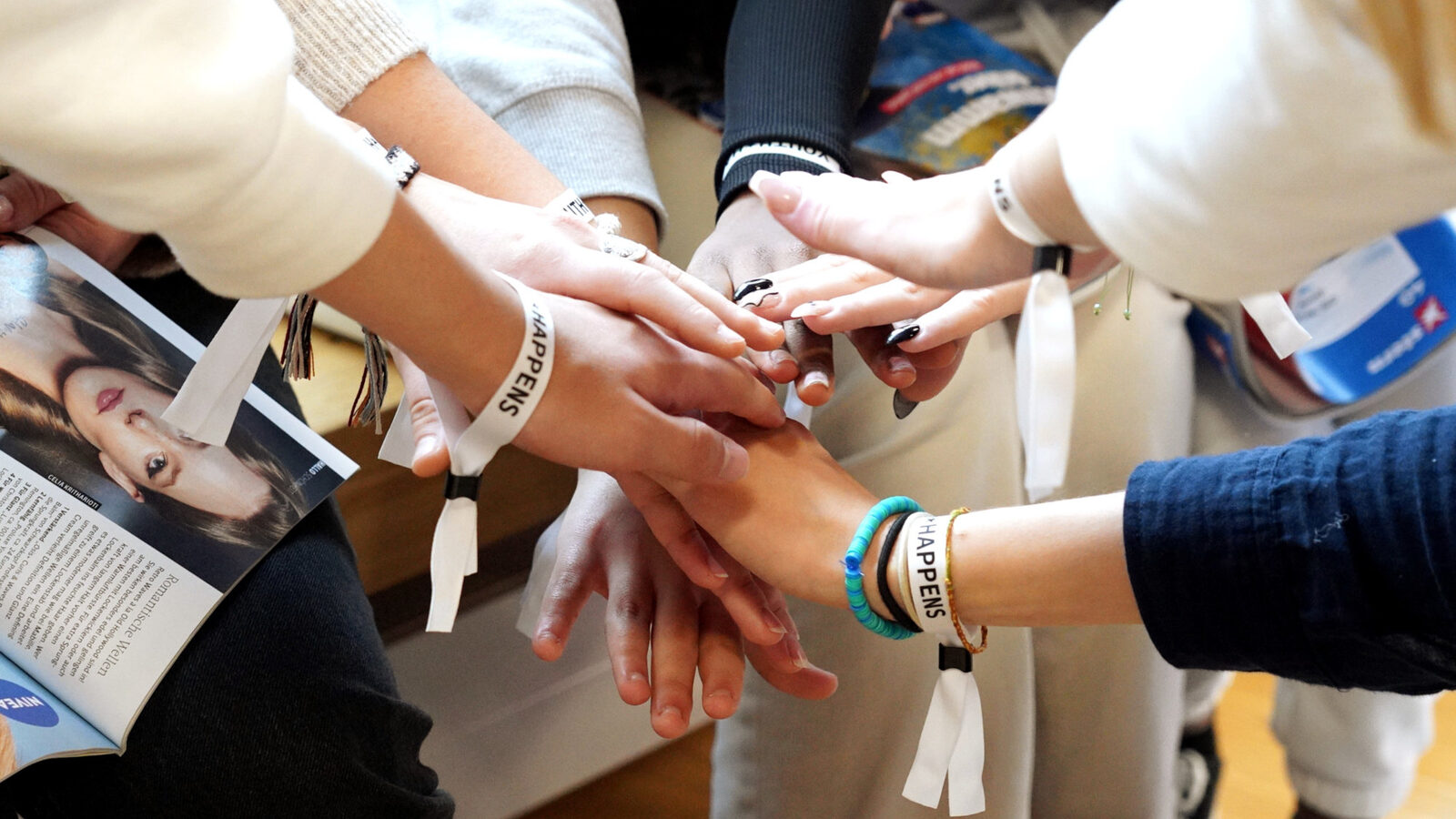 Mehrere Hände von jungen Menschen greifen zusammen in der Mitte. Sie tragen Armbänder, auf ihnen steht "Youth happens".