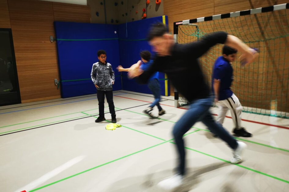 Unscharf sieht man einen jungen Mann einen großen Schritt machen. Er steht in einer Sporthalle vor einem Tor. Andere Kinder sind im Hiintergrund.
