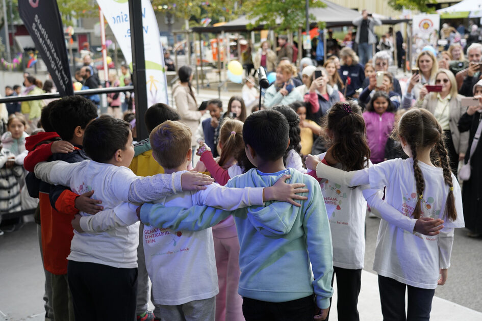 Mehrere Kinder stehen im Halbkreis, sind von hinten zu sehen auf einer Bühne. Sie haben die Arme hintereinander gelegt,