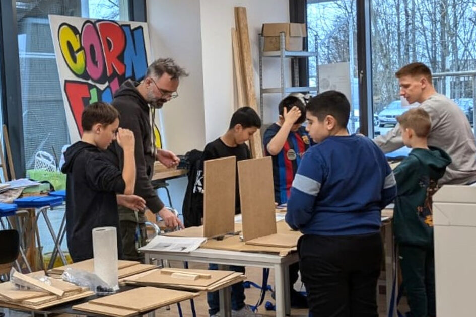 Mehrere kinder und zwei Erwachsene stehen an Tischen mit Holzplatten und werken.