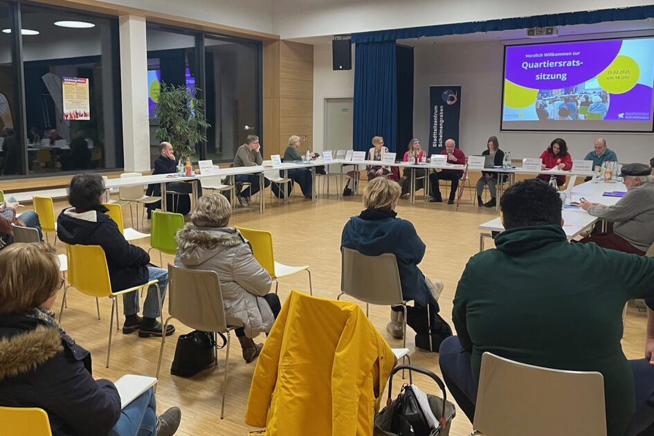 Unter einer Leinwand sitzen an langen Tischen mehrere Menschen. Davor sitzen weitere Menschen auf Stühlen.