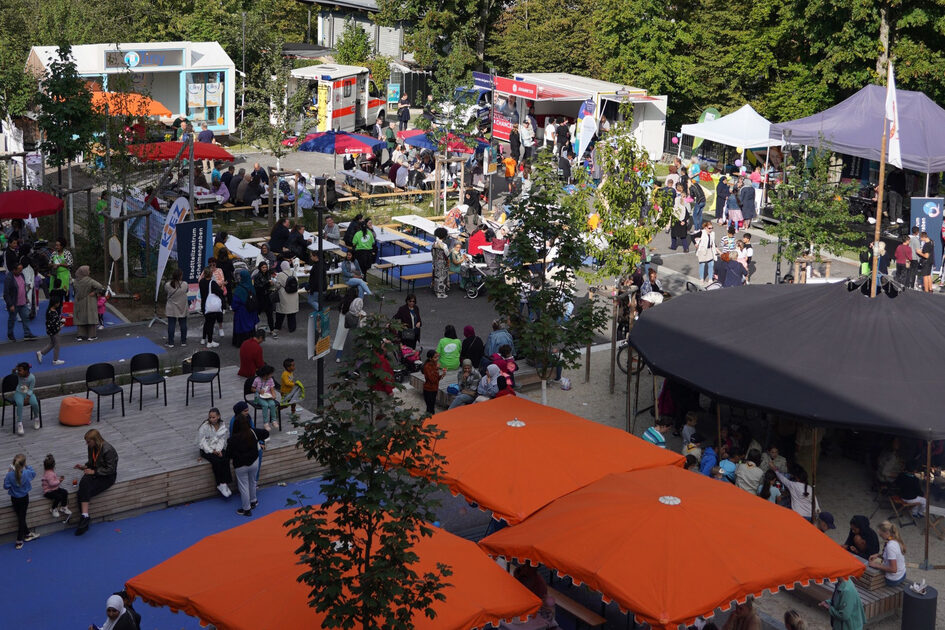 Von oben ist ein Stadtteilfest zu sehen. Mehrere Pavillons und Schrime sind aufgebaut, dazwischen sieht man Menschen.
