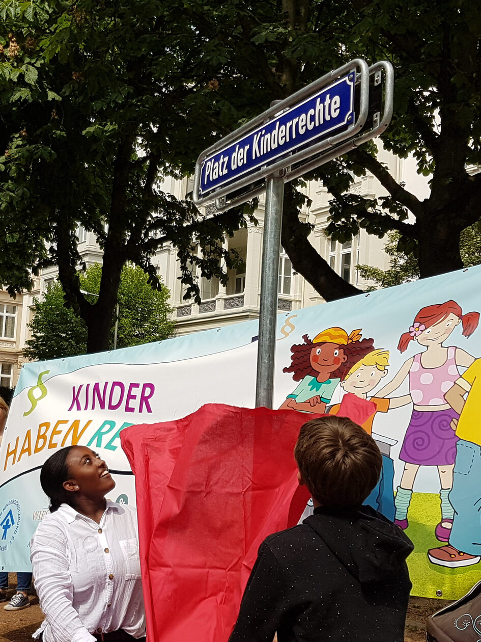 Zwei Jugendliche nehmen eine rote Stoffbahn hinunter, auf einem Schild steht "Platz der Kinderrechte".
