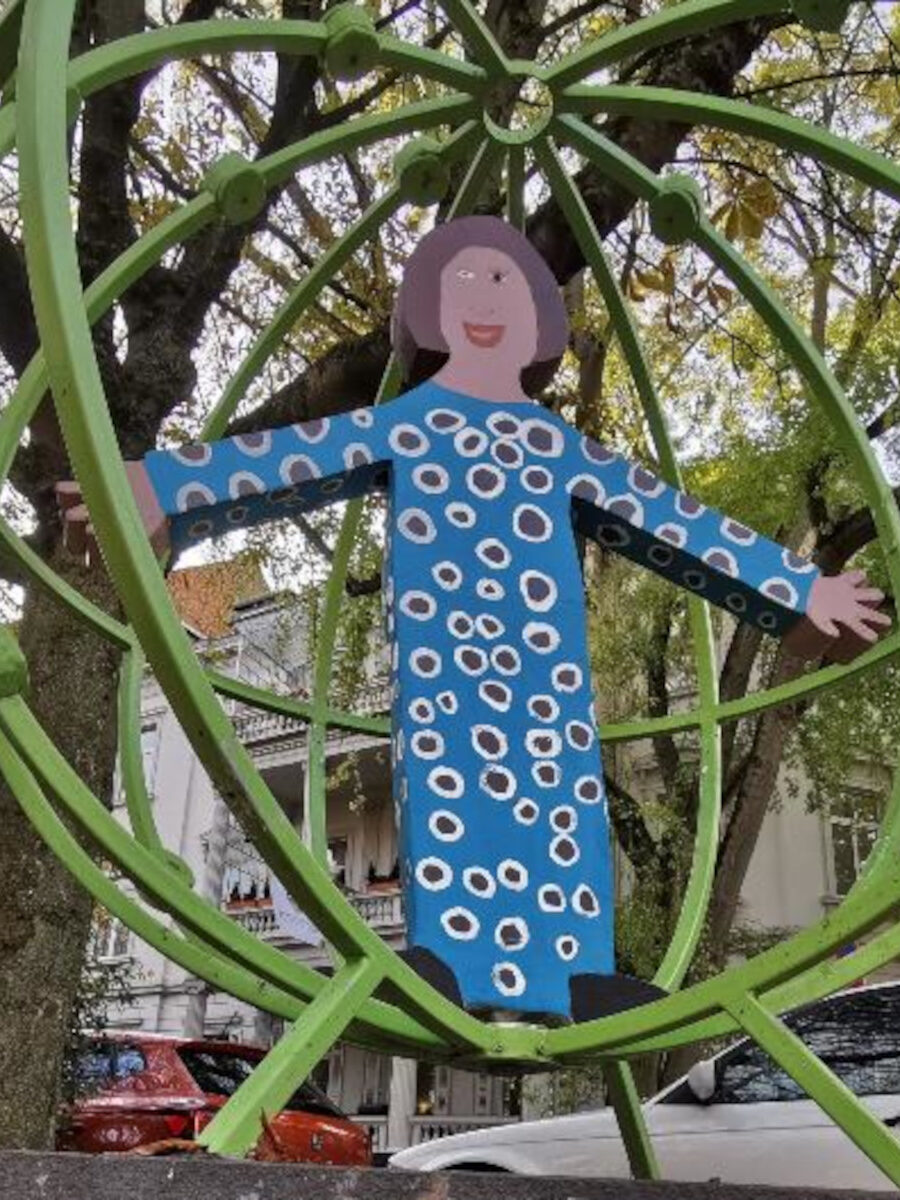 Auf einem städtischen Platz steht in grünen Kreisen eine blau bemalte Frauenfigur aus Holz.