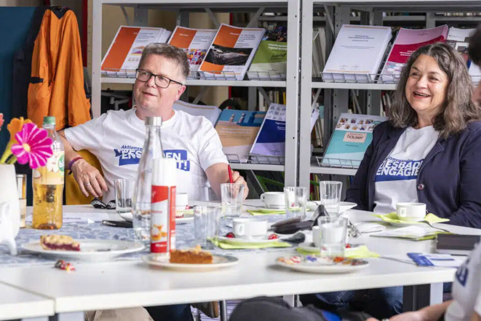 Menschen sitzen um einen Tisch und hören einer weiteren Person zu.