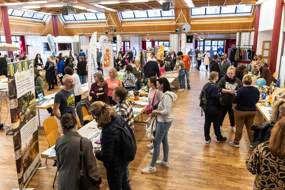 In einer Halle stehen Menschen an Tischen mit Roll-ups und Plakaten. Sie unterhalten sich in kleineren Gruppen.