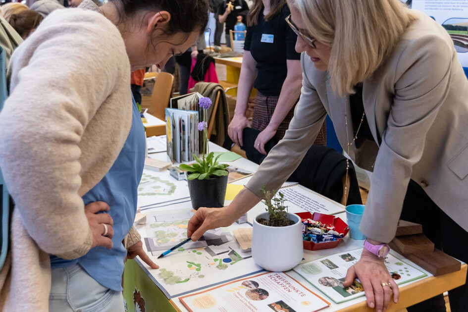Eine Frau steht an einem Tisch und zeigt mit einem Stift auf einen Plan. Eine andere Frau schaut zu.