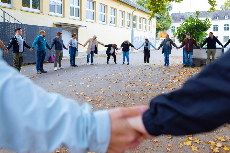 Eine Gruppe Menschen steht im Kreis auf einem Schulhof. Sie halten sich an den Händen.