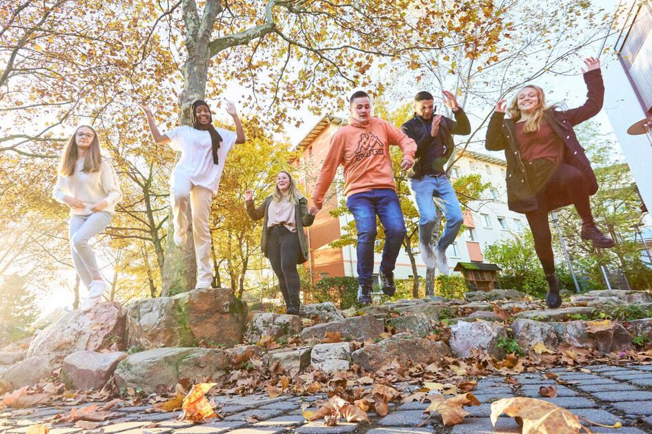 Sechse Jugendliche springen auf einem Schulhof hoch. Blätter auf dem Pflaster zeigen an, dass Herbst ist.